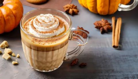 A close-up of a clear glass mug holding a layered Pumpkin Spice Latte. The drink is topped with a generous swirl of whipped cream and a dusting of cinnamon or pumpkin spice. The mug is placed on a dark surface and is accompanied by cinnamon sticks, star anise pods, and miniature pumpkins, evoking an autumnal theme.