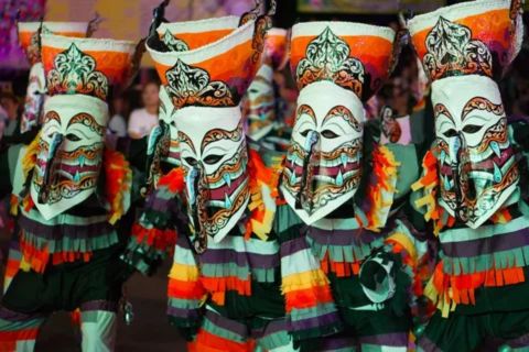 A vibrant, horizontal photograph of the Phi Ta Khon (Ghost Festival) parade in Dan Sai, Loei Province, Thailand. Several participants, mostly men, are walking in a procession on a street lined with spectators. The main focus is on the ghostly, handcrafted masks and colorful patchwork costumes. The masks, known as Phi Ta Khon Lek (Small Ghosts), are grotesque yet whimsical, with tall, exaggerated headpieces made from woven sticky rice baskets and long, pointed wooden noses. They are painted in bright, vibrant colors like red, green, blue, and white. The costumes are made from scraps of brightly colored fabric sewn together in a haphazard style. One ghost in the foreground is shown holding a playful, brightly painted wooden phallus, a traditional part of the fertility-themed ritual. The atmosphere is sunny and festive, with local townspeople and tourists watching the procession from the side of the road. its a one of amazing facts about thailand