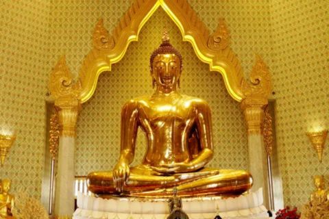 A close-up of a large, brilliant, solid gold seated Buddha statue, possibly the Phra Phuttha Maha Suwan Patimakon (Golden Buddha) from Wat Traimit in Bangkok. The highly polished statue is positioned in the Bhumisparsha Mudra (calling the Earth to witness) on a white pedestal, beneath a decorative, gold-colored archway in a temple interior. The walls are covered in a richly patterned golden wallpaper or mosaic.
