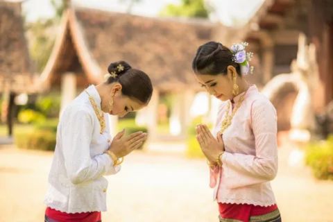 Two Thai women in traditional or formal attire perform the Wai, the customary Thai greeting. They are dressed in delicate white and light pink long-sleeved blouses with high, gold necklaces and bracelets, and red lower garments. They both have their hair styled up and are bowing their heads with their hands pressed together in a prayer-like gesture. They are standing outdoors on a sunny day with traditional Thai wooden architecture visible in the blurred background.