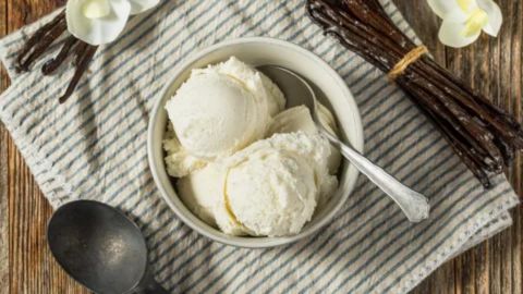An overhead shot of a small white bowl filled with three scoops of creamy, white vanilla ice cream. A silver spoon rests partially in the bowl. The bowl sits on a white and blue striped cloth napkin. Around the bowl are several dark brown vanilla bean pods, some tied in a bundle, and two white vanilla orchid flowers, emphasizing the natural flavor source.