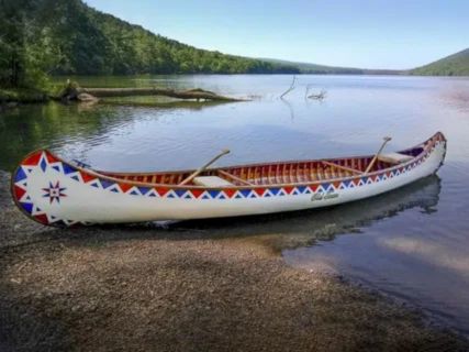 A white wooden canoe with red, white, and blue geometric patterns and a star design rests at the edge of a calm lake. Two paddles are visible inside the canoe, with wooded hills in the distance.