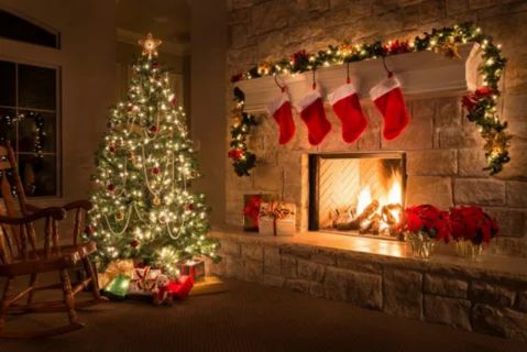Warm, cozy living room decorated for Christmas with a large, lit Christmas tree, stockings hanging above a stone fireplace, and a rocking chair.