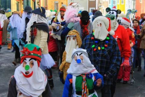 A group of people, dressed in various elaborate disguises with their faces covered, are walking down a street. They are participating in a Mummering parade, a traditional Newfoundland Christmas custom. Costumes include pillowcase-like face coverings, Santa hats, garlands, and mismatched clothing