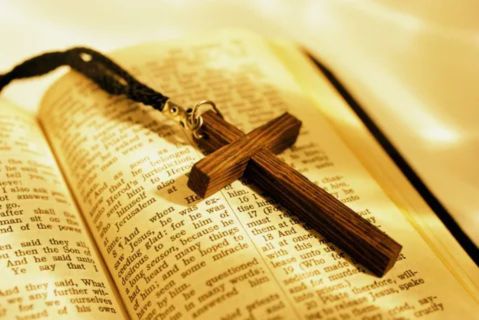 A close-up, warm-toned photograph of a small wooden cross pendant resting on the open pages of a well-worn Bible.