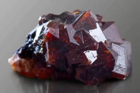 A high-detail macro photograph of a raw, deep red garnet gemstone. The stone exhibits intricate, geometric crystalline facets and rich burgundy tones, set against a smooth, dark grey reflective surface.